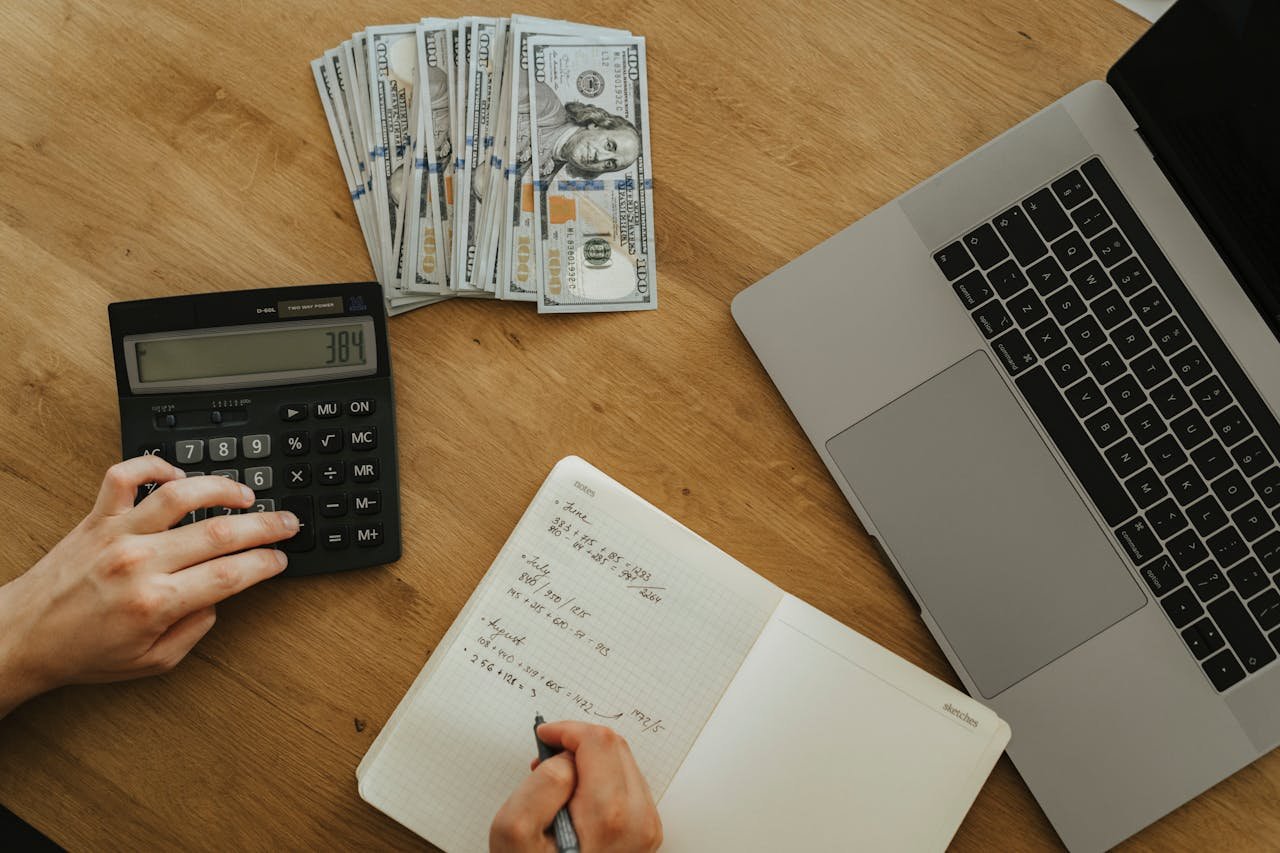 Hands using calculator and notebook, planning finances, with cash and laptop on wooden table.